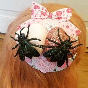 May include: A white and rose gold glitter heart hair clip with black spider accents and pink and white hearts.