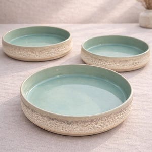 Può includere: Tre piatti da portata in ceramica. Ogni piatto ha un interno turchese chiaro e un esterno strutturato bianco sporco. I piatti sono rotondi e poco profondi, adatti per servire antipasti o piccole porzioni.