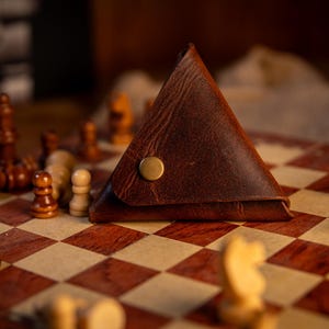 May include: A brown leather triangular coin purse with a gold snap closure rests on a chessboard. Chess pieces in brown and white are arranged around the purse, suggesting a game in progress. The board has a red and tan checkered pattern.