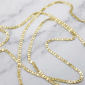 May include: A gold-toned chain necklace featuring a series of flat, elongated, interlocking links. The chain is arranged in a loose, winding pattern, showcasing the polished, reflective surface of the metal links throughout the entire length.