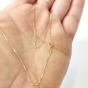 May include: A gold chain necklace with a lobster clasp closure. The chain is a box style and is shown on a hand.