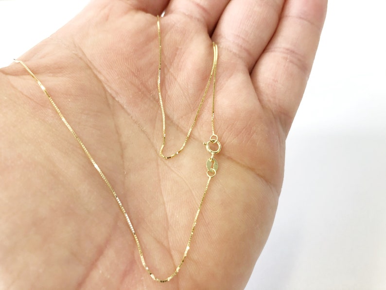 May include: A gold chain necklace with a lobster clasp closure. The chain is a box style and is shown on a hand.