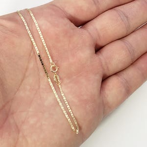May include: A thin gold chain necklace featuring flat, rectangular links connected in a continuous pattern. The necklace includes a small circular spring ring clasp and a tiny metal tag near the closure, showcasing a polished metallic finish.