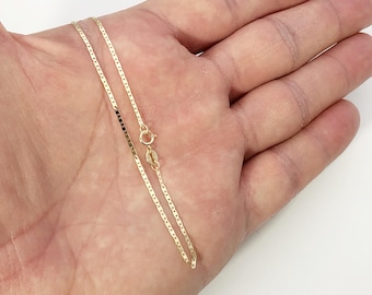 May include: A thin gold chain necklace featuring flat, rectangular links connected in a continuous pattern. The necklace includes a small circular spring ring clasp and a tiny metal tag near the closure, showcasing a polished metallic finish.