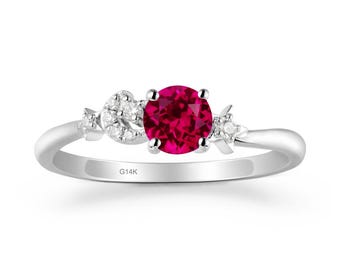 Könnte beinhalten: Ein silberner Ring mit einem großen, runden, leuchtend roten Edelstein im Mittelpunkt. Der Ring ist mit kleineren, klaren Edelsteinen und dekorativen floralen und halbmondförmigen Motiven auf beiden Seiten des Hauptsteins verziert. Das Band ist glatt und poliert, mit der Gravur "G14K".