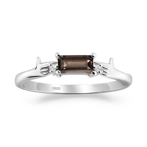 May include: A silver ring featuring a rectangular, smoky brown gemstone at the center, flanked by two smaller, clear gemstones. The ring has a delicate design with stylized hand-like accents on either side of the central stone. The inscription "S925" is visible on the band.