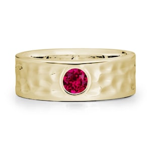 May include: A gold-colored ring with a hammered texture and a round, vibrant red gemstone. The ring's band is thick and features a textured surface. The gemstone is set in a circular bezel.