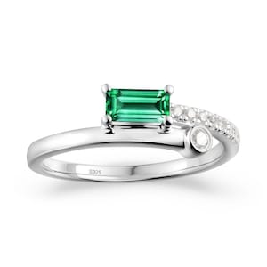 May include: A silver ring featuring a rectangular emerald-cut green gemstone as the focal point. The ring has a delicate, curved band with a row of small, clear stones and a single round stone. The inscription "S925" is visible on the band.