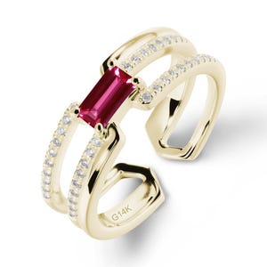 Può includere: Un anello color oro con una gemma rettangolare rosso intenso al centro. L'anello presenta un design a doppia fascia, con piccole pietre trasparenti incastonate lungo le fasce. L'iscrizione "G14K" è visibile sulla fascia dell'anello.
