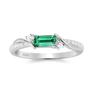 May include: A silver ring featuring a central emerald-green baguette-cut stone flanked by two small round clear stones. The band has a bypass design with additional small clear stones set along the sides and is stamped with S925 on the inner band.