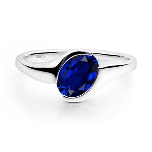 May include: A silver ring featuring a polished band with an oval-cut, deep blue gemstone set in a tension-style mounting. The inner band is clearly engraved with the text S925, indicating the metal composition of this jewelry piece.