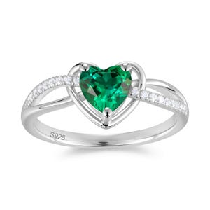 Puede incluir: Un anillo de plata con una piedra preciosa verde esmeralda en forma de corazón. El anillo tiene un diseño delicado con pequeñas piedras claras a lo largo de la banda. La inscripción "S925" es visible.