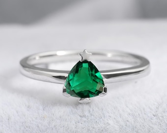 May include: A silver ring featuring a triangular, emerald-green gemstone shaped like a Christmas tree. A small star sits atop the tree. The ring is set on a white surface.