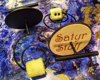 May include: A black metal spinning wheel with two wooden discs. The top disc has a black surface and the bottom disc has the text "Satyr Stuff" engraved on it. The spinning wheel is on a blue and purple background.