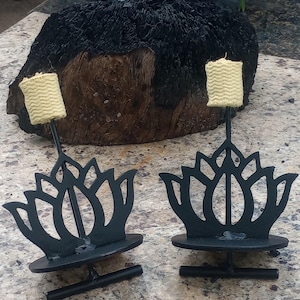 May include: Two black metal lotus flower candle holders with a beige wick. The candle holders have a round base and a decorative lotus flower design. The wicks are rectangular and made of a woven material. The background features a dark wooden log.