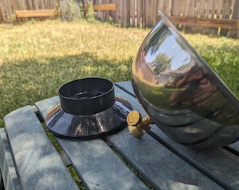 May include: A disassembled, shiny metal outdoor cooking appliance. The components include a large, hemispherical bowl, a black base, and a brass-colored valve. The items are resting on a weathered, blue-green picnic table in a backyard setting.