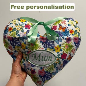 Peut inclure: Coussin en forme de c&oelig;ur avec un motif floral et un ruban vert. Le coussin porte l'inscription brod&eacute;e "Mum". Le fond est blanc avec des fleurs et des papillons color&eacute;s. Le texte "Free personnalisation" est en haut.
