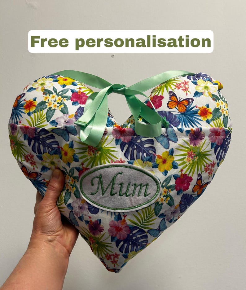 Peut inclure: Coussin en forme de c&oelig;ur avec un motif floral et un ruban vert. Le coussin porte l'inscription brod&eacute;e "Mum". Le fond est blanc avec des fleurs et des papillons color&eacute;s. Le texte "Free personnalisation" est en haut.