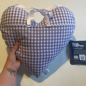 Op de afbeelding: Een hartvormig kussen van paars en wit gingham met een wit satijnen lint eromheen gebonden. Het kussen wordt vastgehouden door een hand. Op de achtergrond is een blauw en wit pakketje te zien met de tekst "Kool Pak".