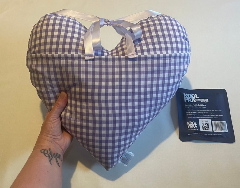 Op de afbeelding: Een hartvormig kussen van paars en wit gingham met een wit satijnen lint eromheen gebonden. Het kussen wordt vastgehouden door een hand. Op de achtergrond is een blauw en wit pakketje te zien met de tekst "Kool Pak".