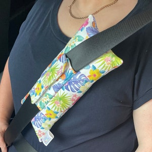 May include: A white seatbelt shoulder pad with a tropical floral print in shades of pink, yellow, green, and blue. The pad is attached to a black seatbelt.
