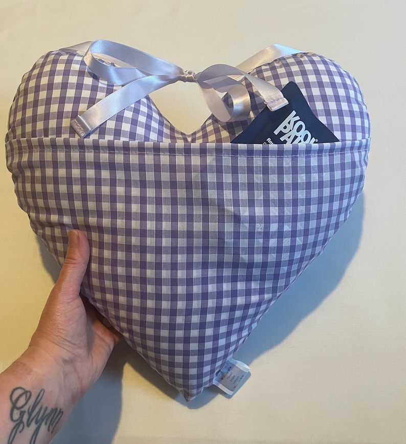 Op de afbeelding: Een hartvormig kussen van paars-wit gingham met een blauwe zak aan de voorkant. De zak heeft een wit label met de tekst "Kool Pad" erop.