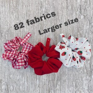 May include: Three fabric scrunchies: red and white gingham, solid red, and white with red strawberries. The image text reads "82 fabrics" and "Larger size". These hair accessories are a larger size.