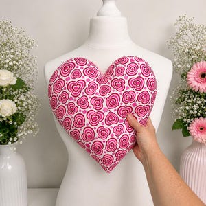 May include: A heart-shaped pillow featuring a repeating pattern of nested pink and magenta hearts on a white background. The pillow is held against a white mannequin torso, showcasing the vibrant heart-themed fabric design and the soft, plush texture.