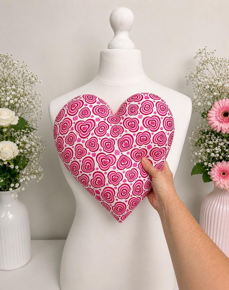 May include: A heart-shaped pillow featuring a repeating pattern of nested pink and magenta hearts on a white background. The pillow is held against a white mannequin torso, showcasing the vibrant heart-themed fabric design and the soft, plush texture.