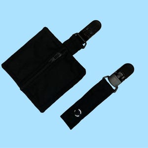 Könnte beinhalten: Zwei schwarze Stoffclips mit einer Reißverschlusstasche. Die Clips haben einen Kunststoffverschluss und ein schwarzes Stoffband. Die Tasche hat einen Reißverschluss.