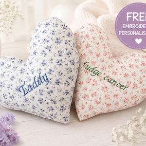 May include: Two heart-shaped pillows with floral patterns. The left pillow is white with blue flowers and the embroidered name Taddy. The right pillow is white with pink flowers and the embroidered text fudge cancer. A purple circle graphic reads FREE EMBROIDERED PERSONALISATION.