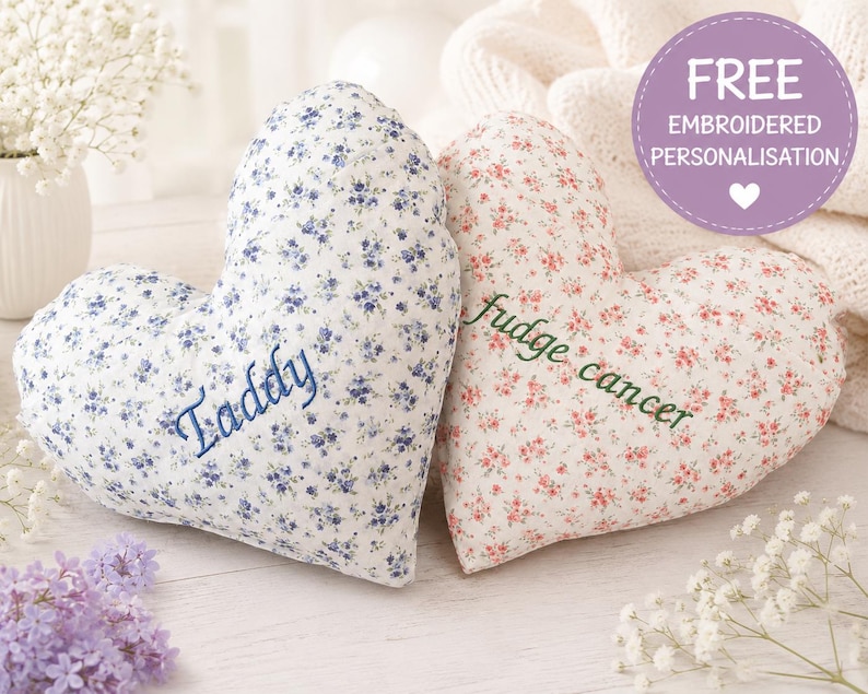 May include: Two heart-shaped pillows with floral patterns. The left pillow is white with blue flowers and the embroidered name Taddy. The right pillow is white with pink flowers and the embroidered text fudge cancer. A purple circle graphic reads FREE EMBROIDERED PERSONALISATION.
