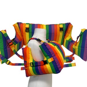 May include: A rainbow striped baby carrier with black buckles and straps. The carrier is displayed on a white mannequin. The carrier has a padded design with multiple sections for support and comfort. The colours include red, orange, yellow, green, blue, and purple.