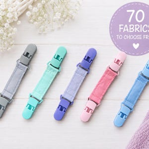 以下が含まれることがあります： グレー、ミントグリーン、ラベンダー、ピンク、ライトブルーのプラスチック製クリップが付いた布製おしゃぶりホルダー5本。右上の紫色の円形グラフィックには、70 FABRICS TO CHOOSE FROMというテキストと小さな白いハートのアイコンが表示されています。