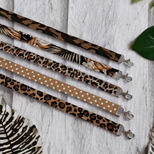 Könnte beinhalten: Fünf Stoff-Lanyards mit Metall-Karabinern. Die Lanyards weisen verschiedene Tiermuster auf, darunter Camouflage, Leopardenflecken und ein beiges Polka-Dot-Design. Die Lanyards sind diagonal vor einem weißen Holzmaserungshintergrund angeordnet.