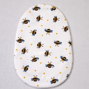 May include: White, oval fabric featuring a repeating pattern of cartoon bees in brown, blue, and yellow, with small yellow dots. The design suggests a baby item.