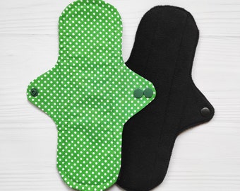 May include: Two reusable cloth menstrual pads. One pad features a bright green fabric with a white polka dot pattern and two green plastic snap closures on the wings. The second pad is made of a solid black fabric with a single black snap closure.