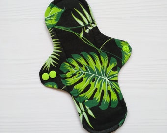 May include: A reusable cloth menstrual pad featuring a black background with a vibrant green monstera leaf and tropical foliage pattern. The pad has a contoured shape with two bright lime green plastic snap buttons on the side wings.
