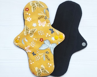 May include: Two reusable cloth menstrual pads. One pad features a mustard yellow fabric with a pattern of white and light blue butterflies, small flowers, and sprigs, secured with yellow snaps. The other pad is solid black with a grey snap.