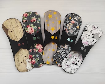 May include: Five reusable cloth menstrual pads with black fabric wings and snap closures. The patterns include beige bears, dark floral roses, yellow stars on gray, dark floral foliage, and white cats with glasses on a black and white background.