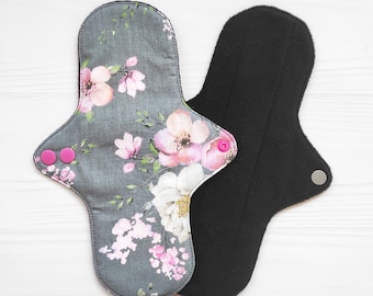 May include: Two reusable cloth menstrual pads. One pad features a dark gray fabric with a floral pattern in shades of pink and white, secured with two bright pink plastic snaps. The second pad is solid black with a single silver-colored metal snap.