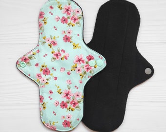 May include: Two reusable cloth menstrual pads. One pad features a light mint green fabric with a pink and yellow floral pattern and two mint green plastic snap buttons. The other pad is solid black with a single silver-colored metal snap button.