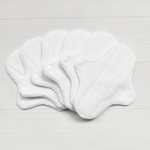 Puede incluir: Cinco compresas menstruales de tela reutilizables blancas con cierres a presión. Las compresas están dobladas y apiladas sobre una superficie de madera blanca.