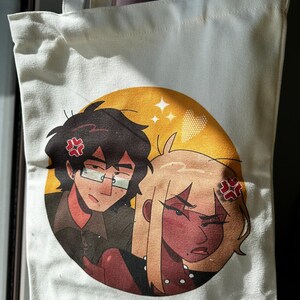 Puede incluir: Bolso tote de lona blanca con una colorida ilustración de dos personajes de estilo anime con expresiones de enfado. La obra de arte está ambientada sobre un círculo amarillo con detalles de estrellas. Los personajes tienen pelo oscuro y gafas, y pelo rubio.