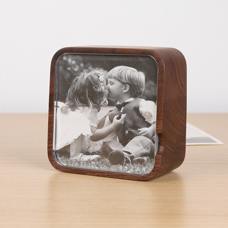 May include: A wooden photo frame with a clear glass insert. The frame features a black and white photo of two children kissing. The frame is on a wooden surface.