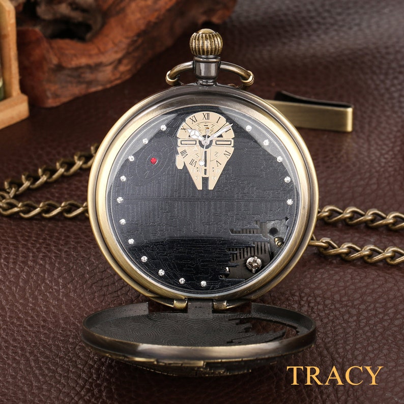 May include: A vintage-style pocket watch with a black face and a silver chain. The watch face features a detailed design of the Millennium Falcon spaceship from Star Wars. The watch has a gold-tone finish and a clear glass cover.