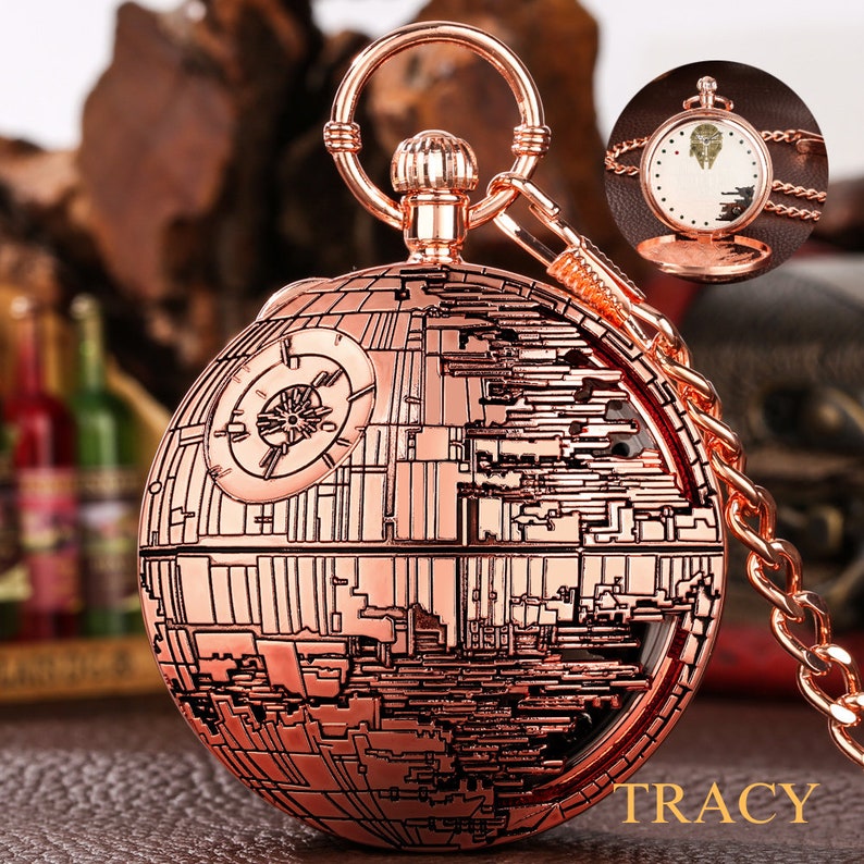 May include: A rose gold pocket watch with a detailed design of the Death Star from Star Wars. The watch has a chain attached to it and the face of the watch is visible in the background.