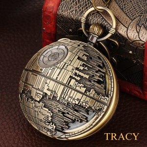 May include: A gold-toned pocket watch with a detailed design of the Death Star from Star Wars. The watch has a black face and a chain attached to the top.