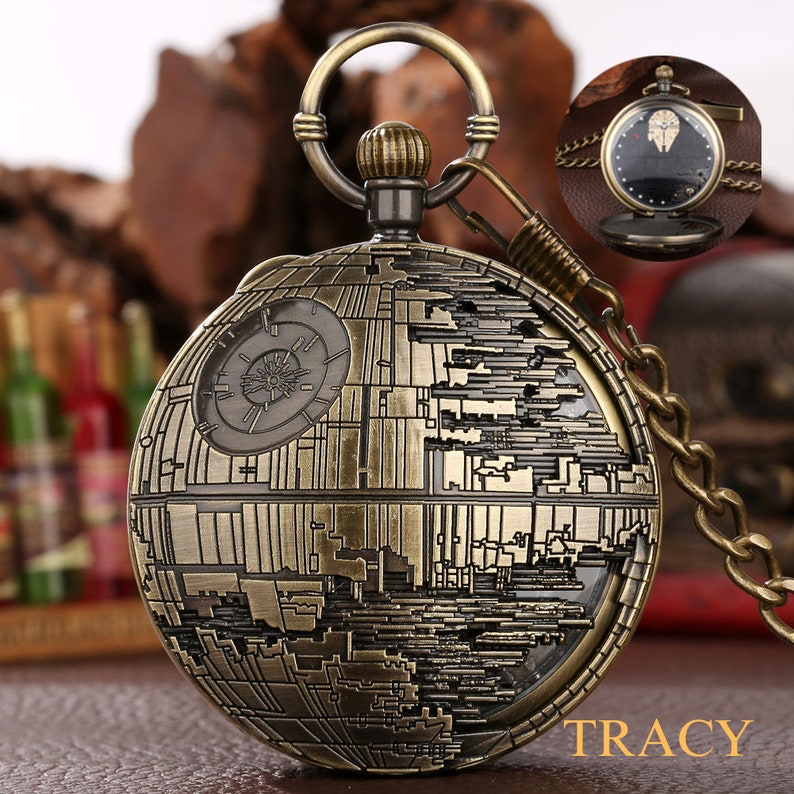 May include: A gold-toned pocket watch with a chain. The watch face features a detailed design of the Death Star from Star Wars. The watch has a round, slightly domed face and a small, round knob on the top.