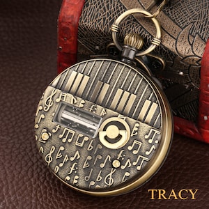 May include: A gold-toned pocket watch with a musical theme. The watch face features a piano keyboard design and musical notes. The watch has a clear window revealing a small, intricate mechanism.